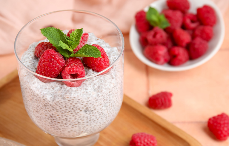 Glas mit Chia‑Pudding, belegt mit frischen Himbeeren und einem Minzblatt, daneben eine Schale mit weiteren Himbeeren.