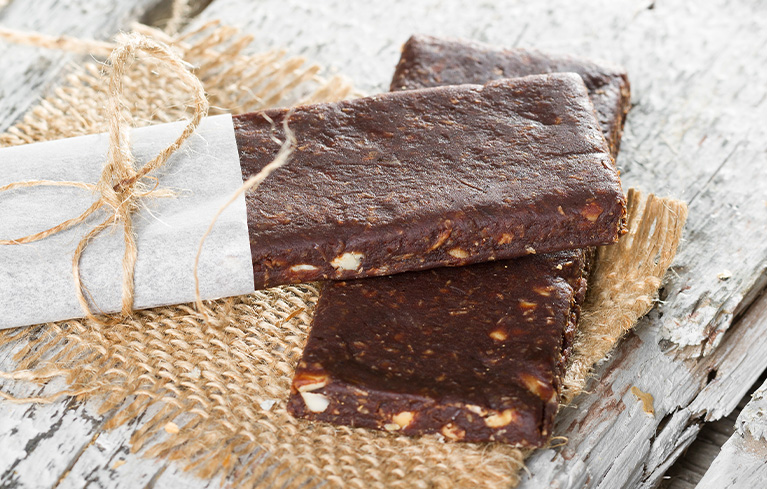 Zwei dunkle Müsliriegel mit Nussstücken, einer davon in Papier und Schnur eingewickelt, auf einer rustikalen Holzunterlage.