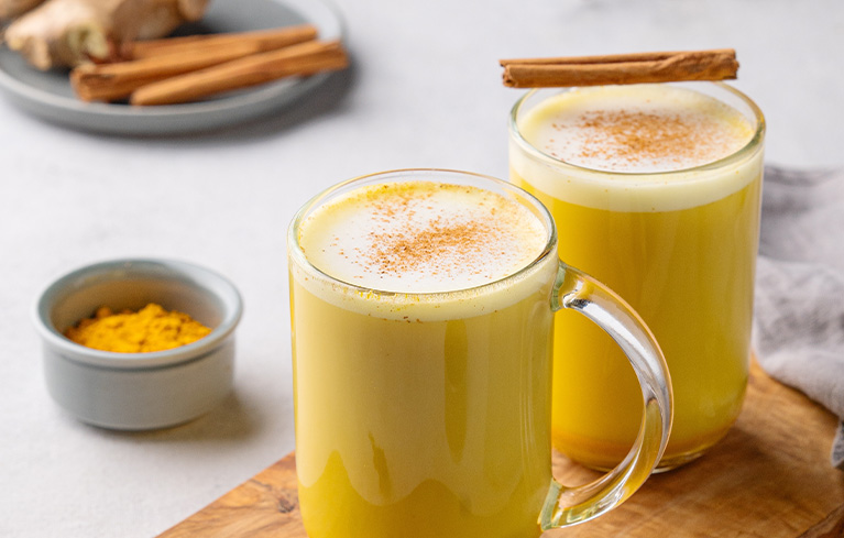 Zwei Gläser Goldene Milch mit Zimtstangen und etwas Zimtpulver darauf, auf einem Holzbrett.