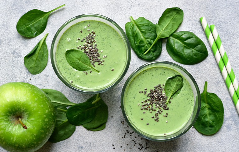 Zwei Gläser grüner Smoothie mit Spinatblättern und Chiasamen, daneben frischer Spinat und ein grüner Apfel auf einer hellen Oberfläche.
