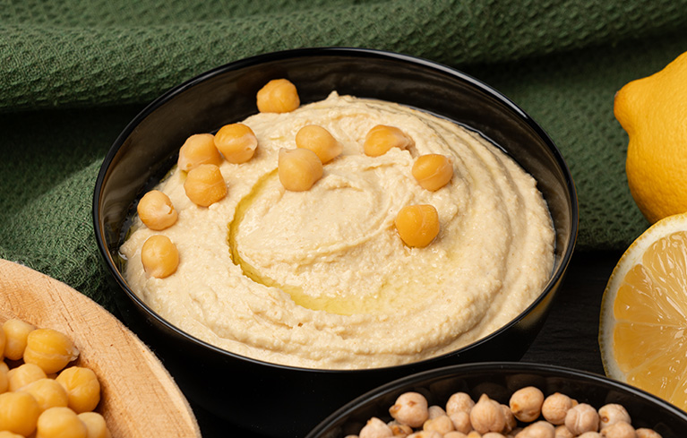 Schale mit Hummus, garniert mit Kichererbsen, daneben weitere Kichererbsen und eine halbierte Zitrone.