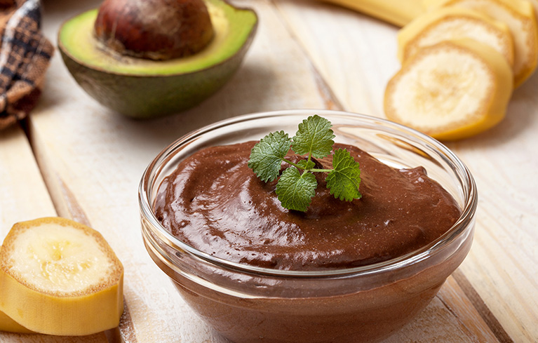 Glasschale mit Schokoladenpudding, garniert mit einem grünen Kräuterblatt, daneben Bananenscheiben und eine halbierte Avocado.