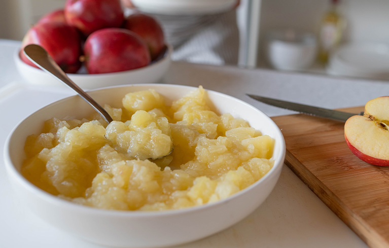 Schüssel mit Apfelmus und einem Löffel darin, daneben ein angeschnittenes Apfelstück und mehrere rote Äpfel im Hintergrund.