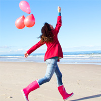 Junge Frau in roten Gummistiefeln und roter Jacke rennt mit Luftballons am Strand