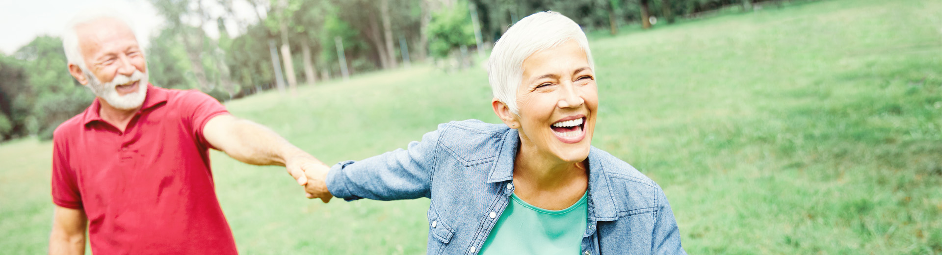 Ältere Frau zieht im Grünen ihren Mann hinter sich her, beide lachen
