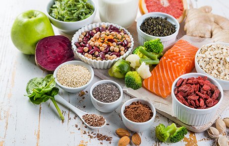 Verschiedene Lebensmittel wie Gemüse, Fisch, Nüsse, Samen und Hülsenfrüchte liegen auf einer hellen Fläche