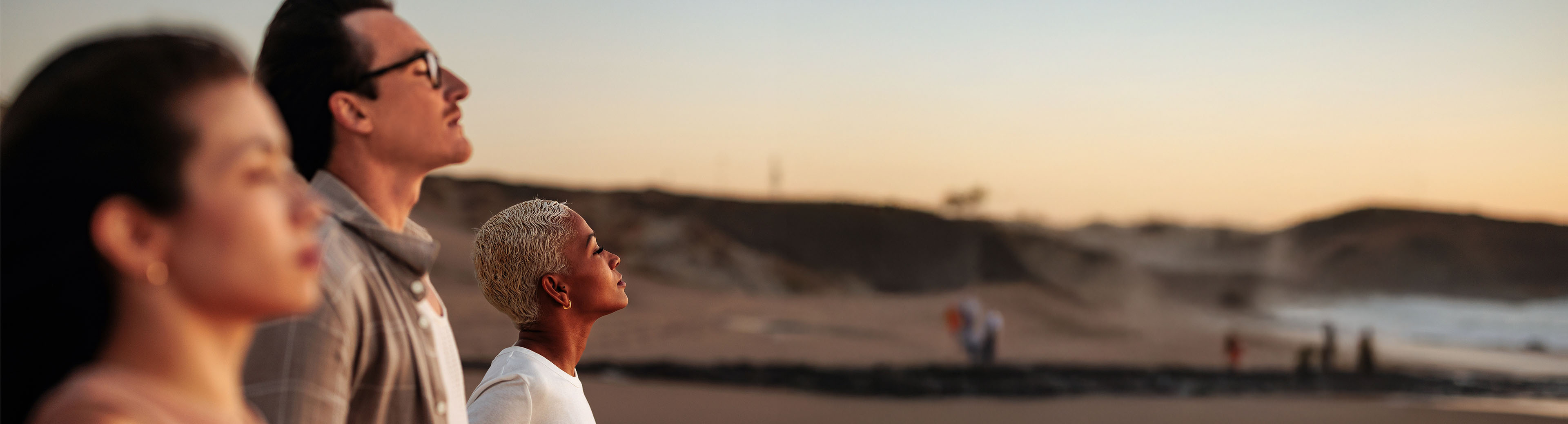 Drei Personen stehen seitlich zum Betrachter am Strand und atmen tief ein, während im Hintergrund die Wellen an die Küste rollen und der Himmel in warmen Farben des Sonnenuntergangs leuchtet.