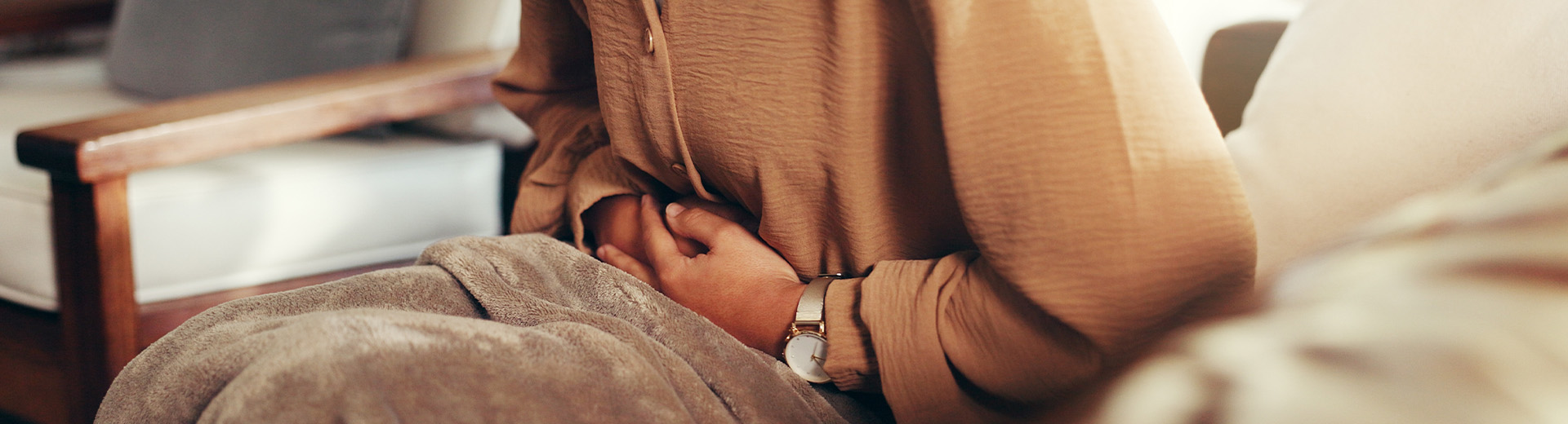 Frau auf Sofa sitzend und in Decke gekuschelt hält sich vor Schmerzen den Bauch