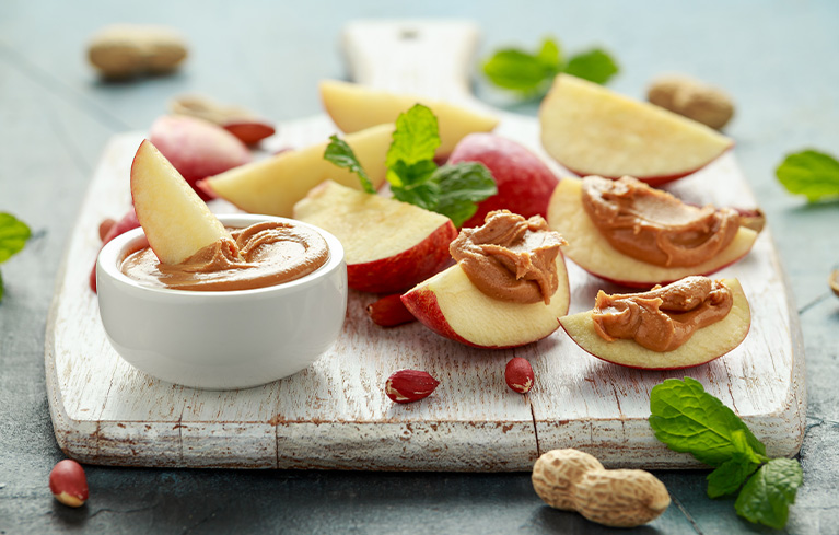 Apfelspalten mit Nussmus auf einem Holzbrett, daneben ein kleines Schälchen mit Nussmus.