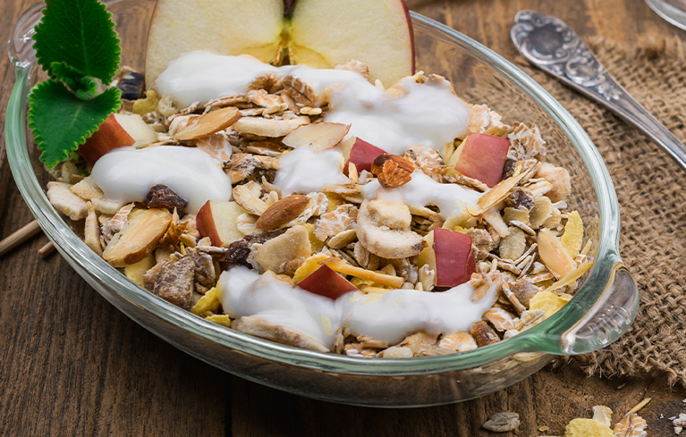 Glasschüssel mit Müsli, Apfelstücken, Nüssen und etwas Joghurt, im Hintergrund eine aufgeschnittene Apfelhälfte.