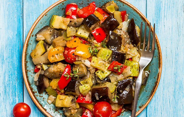 Teller mit gemischtem Gemüse aus Paprika, Zucchini, Aubergine, Tomaten und Zwiebeln, daneben eine Gabel.