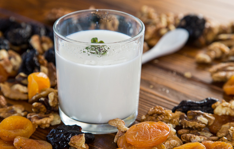 Glas mit Joghurt auf einem Holztisch, umgeben von getrockneten Aprikosen, Walnüssen und Pflaumen.