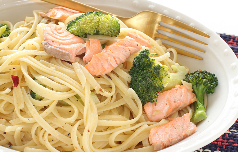 Pasta mit Lachsstücken und Brokkoli auf einem weißen Teller, daneben eine goldene Gabel.