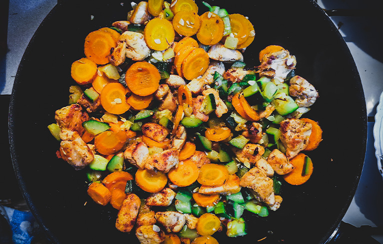 Pfanne mit gebratenem Hähnchen, Karottenscheiben und Gurkenstücken.