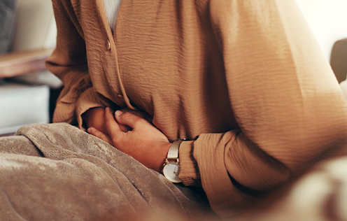 Person sitzt und hält sich mit beiden Händen den Bauch, mit einer Decke zugedeckt.