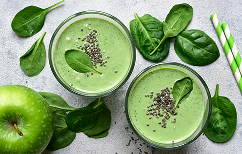 Zwei Gläser grüner Smoothie mit Spinatblättern und Chiasamen, daneben frischer Spinat und ein grüner Apfel auf einer hellen Oberfläche.